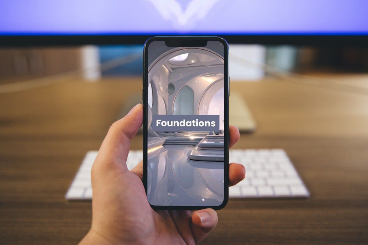 Hand holding a smartphone with 'Foundations' course displayed, in front of a blurred office desk.