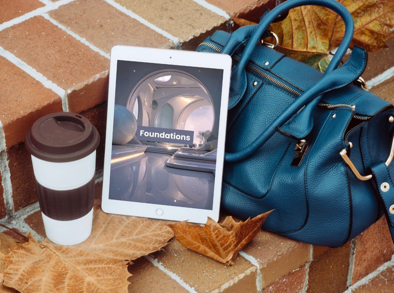 Blue handbag, tablet displaying 'Foundations', and coffee cup on a brick surface with leaves.