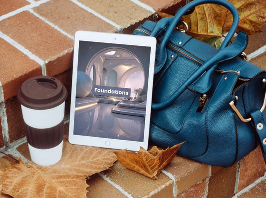 Blue handbag, tablet displaying 'Foundations', and coffee cup on a brick surface with leaves.