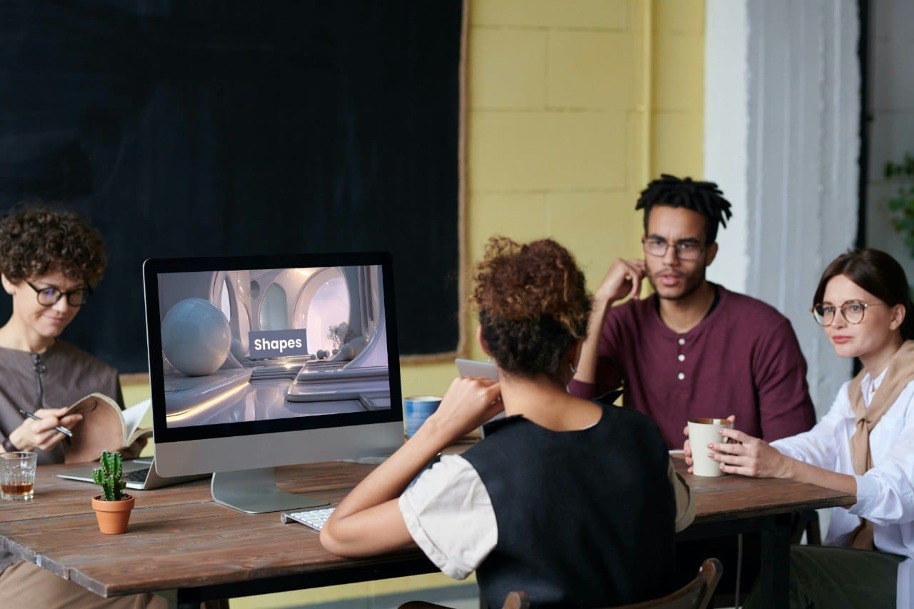 Group of people sitting around a table with a computer screen displaying educational content.