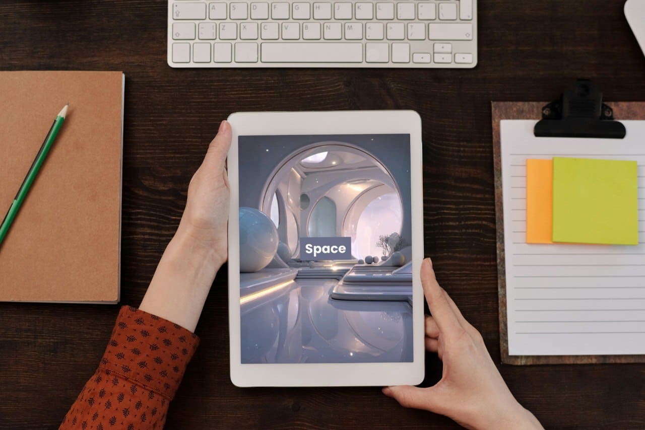 Person holding a tablet with a 'Space' course on a desk with a keyboard, notebook, and post-it notes.